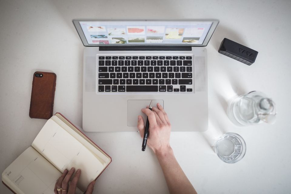 Hender som skisserer i en notatbok ved siden av en laptop